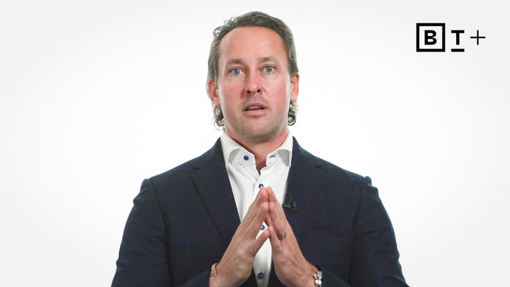 A man in a suit sits against a white background with a BT+ logo, holding his hands together in front of him.