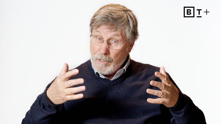 An older man with gray hair and a beard, wearing glasses and a dark sweater, gestures with both hands while speaking in front of a plain white background.