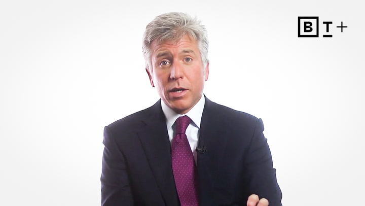 A man in a dark suit and red tie speaks in front of a plain white background with the B+ logo in the top right corner.