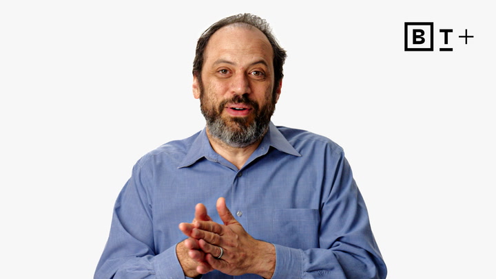 A man with a beard in a blue shirt speaks while gesturing with his hands. A "B T +" logo is visible in the top right corner on a plain white background.