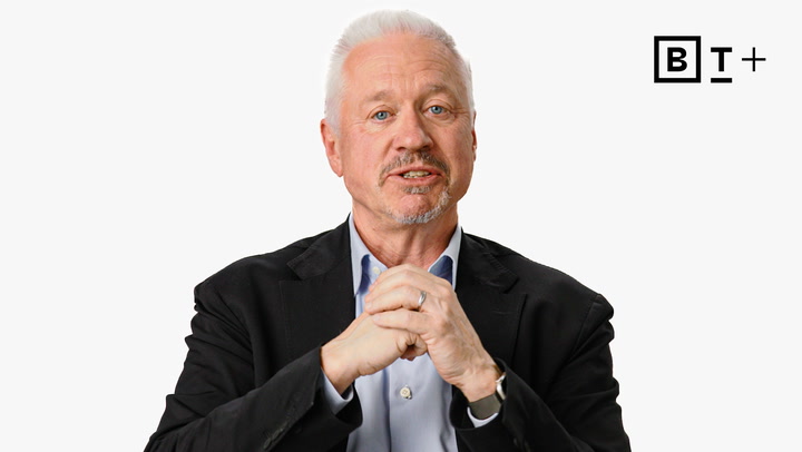A man with white hair and a beard, wearing a dark suit and blue shirt, sits with hands clasped. A "BT+" logo appears in the top right corner on a white background.