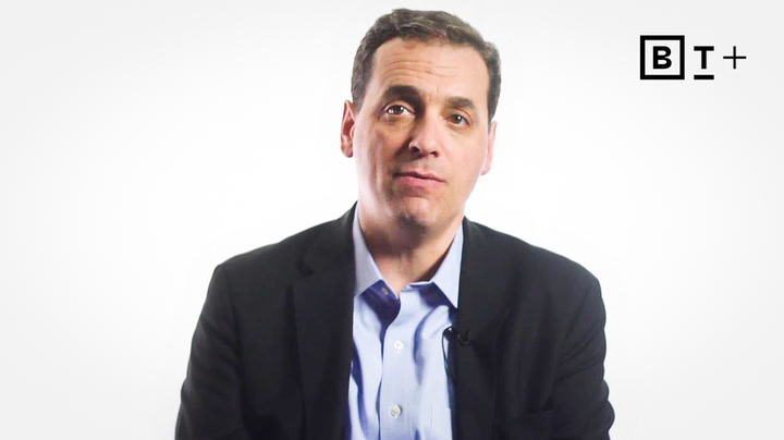 A man in a dark blazer and light blue shirt sits facing the camera against a plain white background with a "BT+" logo in the top right corner.