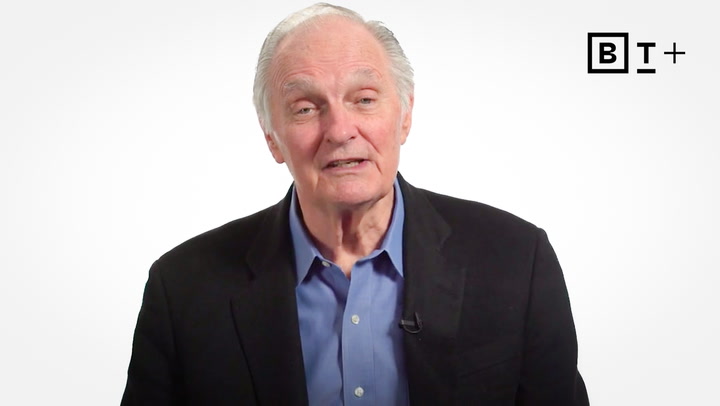 An older man in a dark blazer and blue shirt speaks in front of a white background with a "B T +" logo in the top right corner.