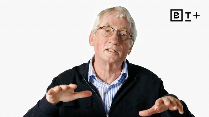 An older man with glasses gestures with both hands while speaking. He wears a dark jacket over a striped shirt. A stylized "B T +" logo appears in the top right corner.