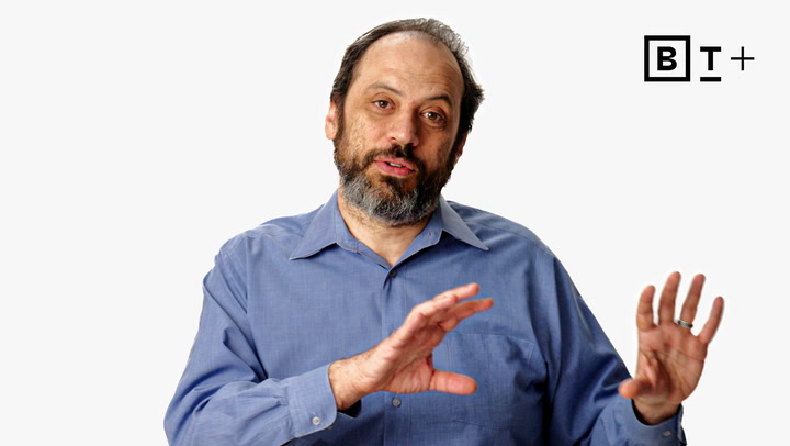 A man with a beard and mustache in a blue shirt gestures with his hands while speaking, with a "B T +" logo in the top right corner on a plain white background.