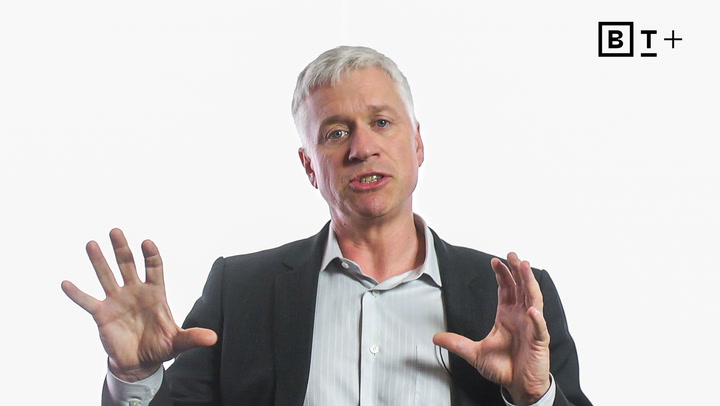 A man in a suit speaks while gesturing with both hands, seated against a plain white background with a "BT+" logo in the top right corner.