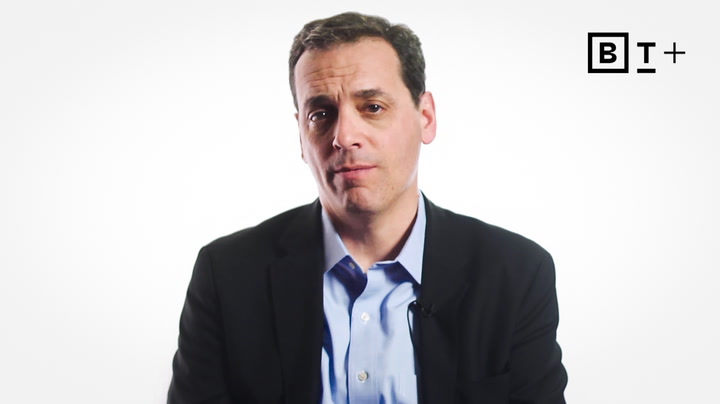 A man in a suit jacket and blue shirt sits against a plain white background with a "B T +" logo in the top right corner.