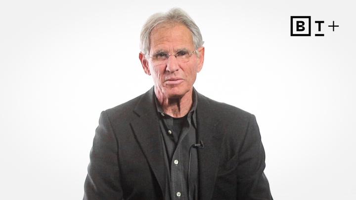 An older man wearing glasses and a dark blazer sits in front of a white background with a "BT+" logo in the top right corner.