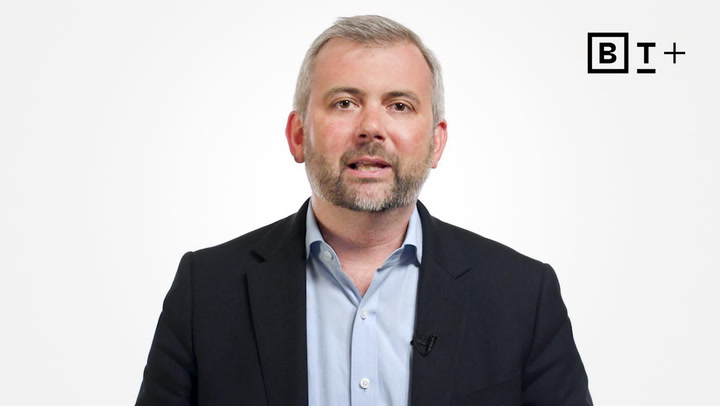 A man in a suit and light blue shirt speaks in front of a plain white background with a "B T +" logo in the top right corner.