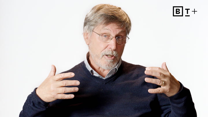 An older man with gray hair and glasses, wearing a dark sweater over a collared shirt, gestures with both hands while speaking in front of a plain white background.