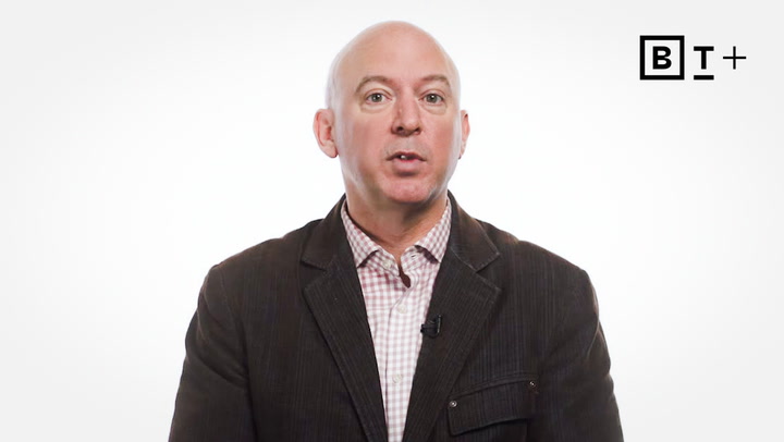 Bald man in a dark jacket and checkered shirt speaks in front of a white background with a "B T +" logo in the top right corner.