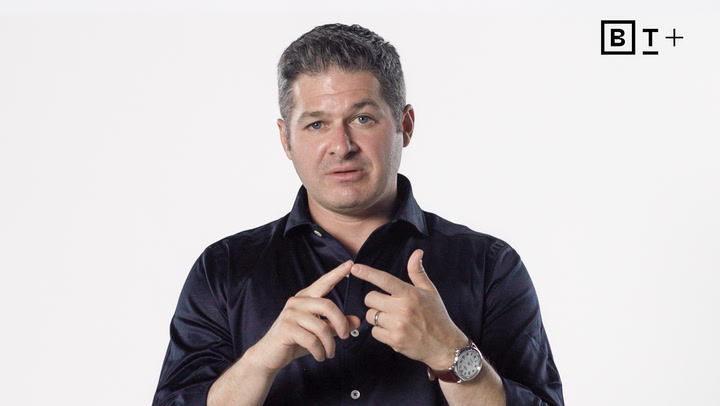A man in a dark shirt gestures with his hands against a plain white background with a "BT+" logo in the top right corner.
