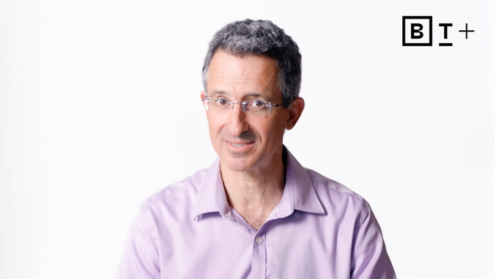 A man with short, curly gray hair and glasses wearing a light purple shirt, sits in front of a plain white background with a black "B T +" logo in the top right corner.