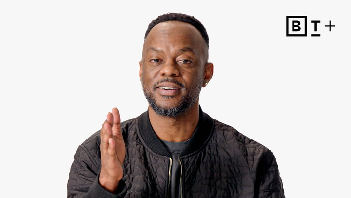 A man with short hair and a beard, wearing a dark quilted jacket, speaks while gesturing with his right hand. A “BT+” logo appears in the top right corner on a white background.