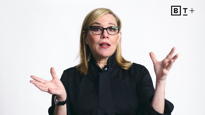 A woman with blond hair and glasses, wearing a black shirt, gestures with both hands while speaking in front of a plain white background.