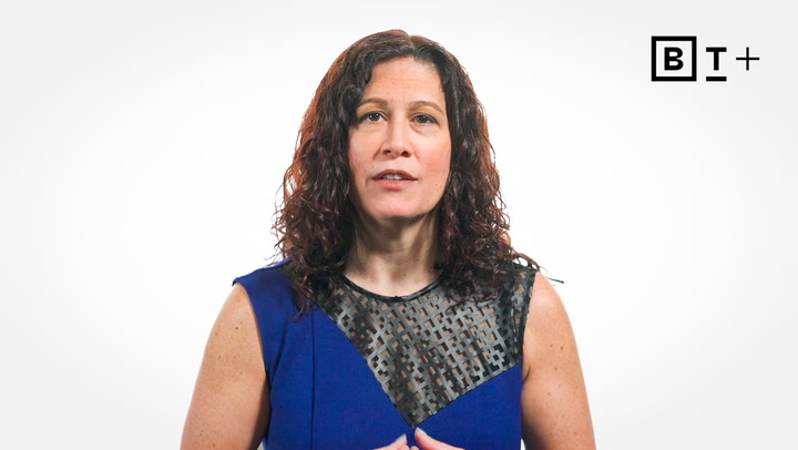 A woman with curly brown hair wearing a blue dress with a black mesh panel stands against a plain white background with a "BT+" logo in the top right corner.
