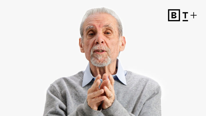 An older man with gray hair and a beard, wearing a light blue collared shirt and gray sweater, gestures with his hands. A "B T +" logo appears in the top right corner.
