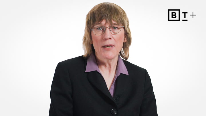 A person with shoulder-length light brown hair, glasses, and a black blazer over a purple shirt sits in front of a white background with a "B T +" logo in the top right corner.