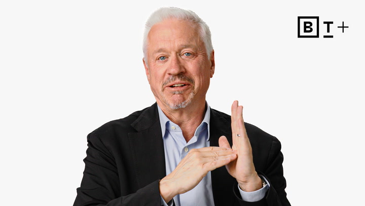 A man with gray hair and a beard, wearing a black blazer and light blue shirt, gestures with his hands in front of a plain white background with a "B T +" logo in the corner.
