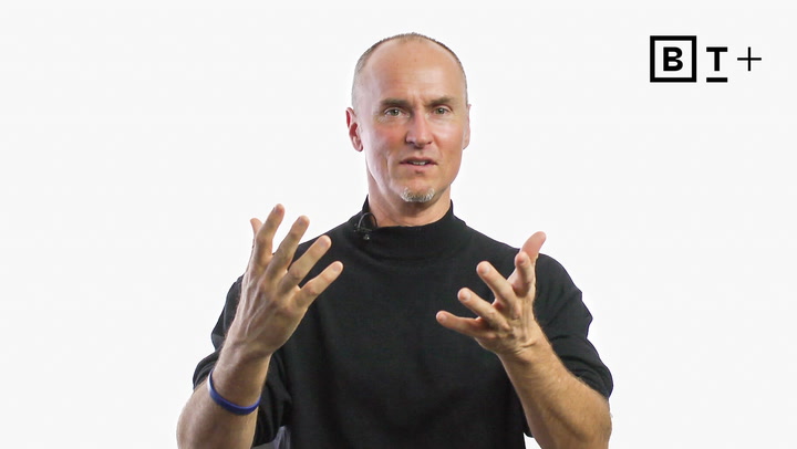 A man in a black turtleneck gestures with both hands while speaking, with a microphone attached to his collar and a logo in the top right corner on a plain white background.