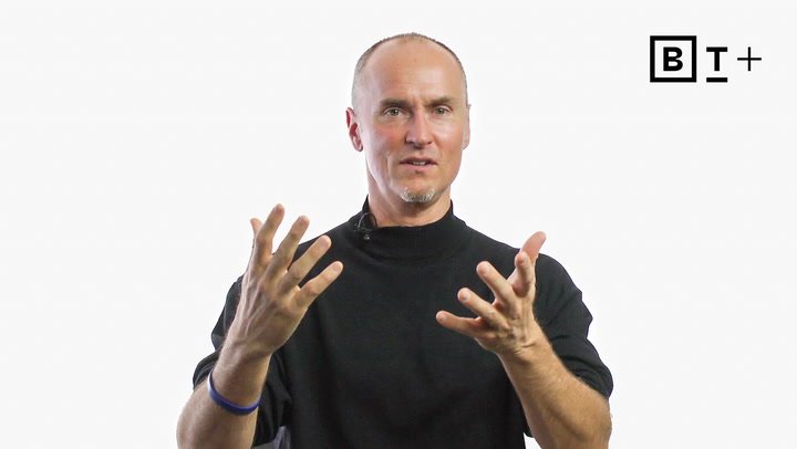 A man in a black turtleneck gestures with both hands while speaking, with a microphone attached to his collar and a logo in the top right corner on a plain white background.