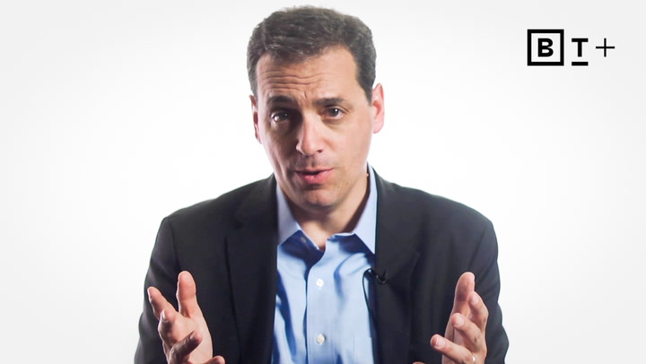 A man in a suit gestures with his hands while speaking in front of a white background with a "BT+" logo in the corner.