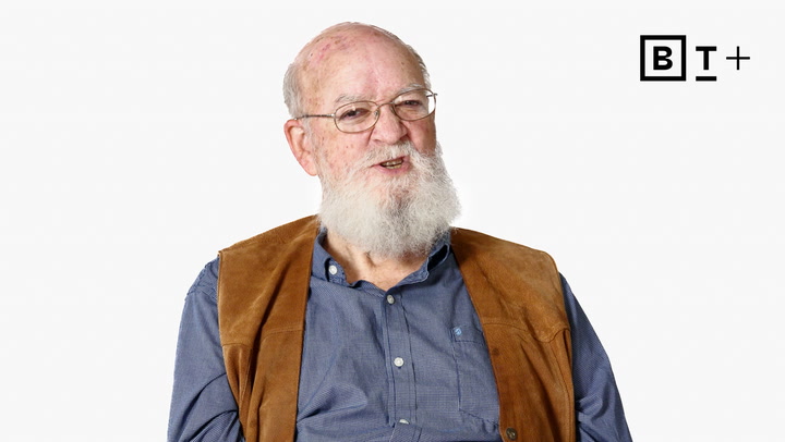 An older man with a white beard and glasses wears a blue shirt and brown vest, seated against a plain white background with a "BT+" logo in the top right corner.