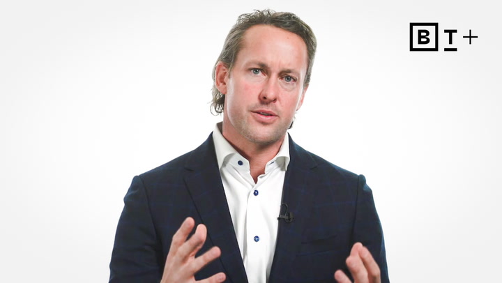 A man in a dark suit and white shirt gestures with his hands while speaking against a plain white background with a B T + logo in the top right corner.