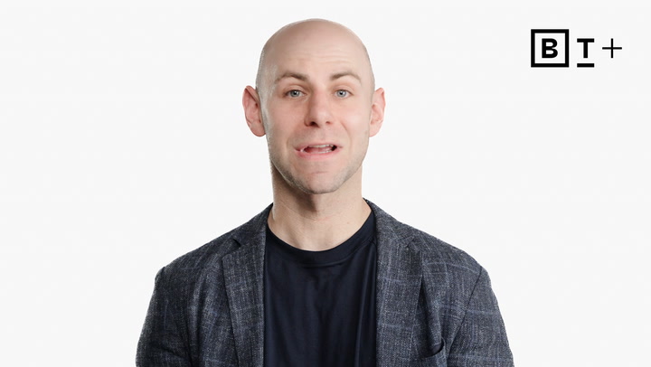 A bald man wearing a dark blazer and shirt speaks in front of a plain white background with a "B T +" logo in the top right corner.
