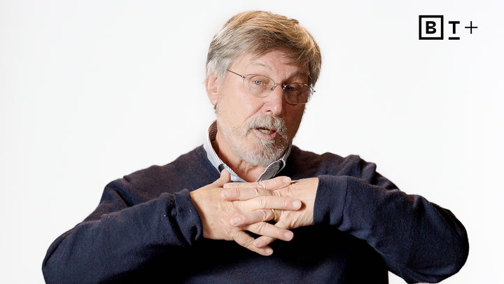 An older man with gray hair and glasses sits against a white background, clasping his hands together and speaking. A logo with the letters "BT+" appears in the top right corner.