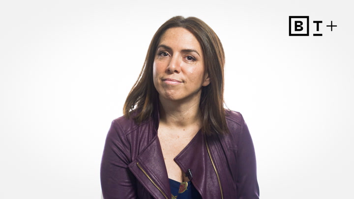 A woman with straight brown hair wearing a purple jacket sits facing the camera against a plain white background with a "BT+" logo in the top right corner.