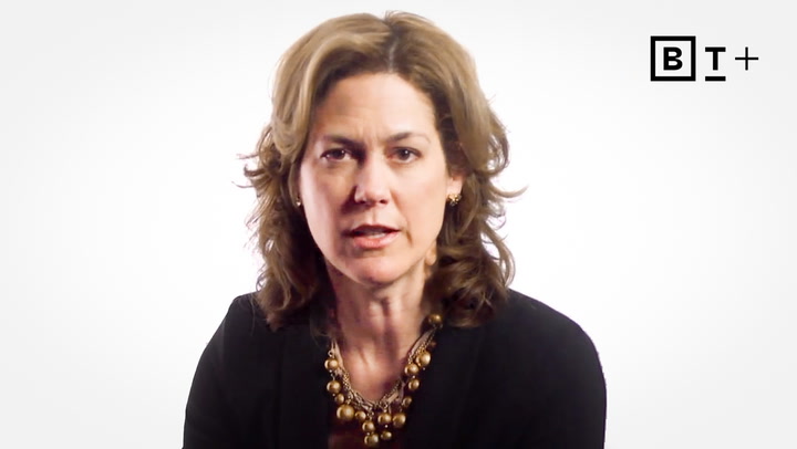 A woman with wavy brown hair wearing a black blazer and a gold necklace sits in front of a white background with a "BT+" logo in the top right corner.