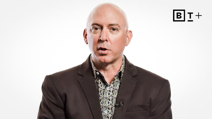 A bald man in a dark blazer and patterned shirt speaks in front of a plain white background with a "B T +" logo in the top right corner.