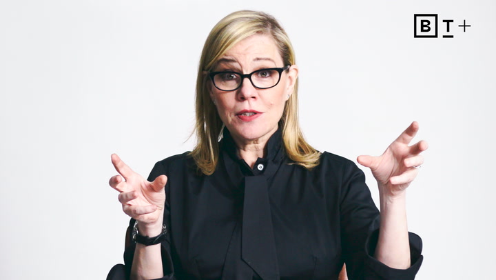 A woman with blonde hair and glasses, dressed in black, gestures with both hands while speaking in front of a plain white background. The letters "BT+" are in the top right corner.