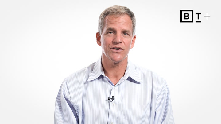A man in a light striped shirt speaks while facing the camera, with a clip-on microphone attached to his collar and a "BT+" logo in the top right corner.