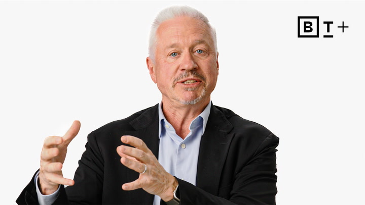 A man with gray hair and a beard, wearing a black blazer and blue shirt, gestures with his hands while speaking. The letters "BT+" appear in the top right corner.