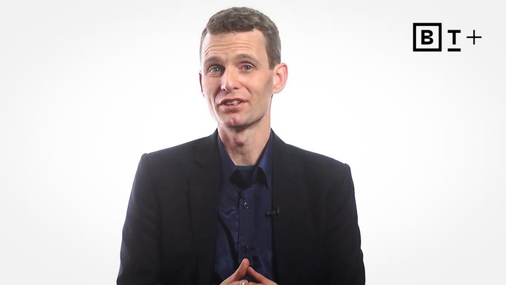 A man in a dark blazer and blue shirt sits in front of a plain white background with a "B T +" logo in the top right corner.