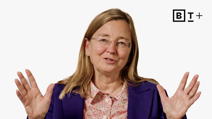 A woman with long hair and glasses speaks while gesturing with both hands, wearing a purple blazer and floral shirt, with a "B T +" logo in the top right corner.