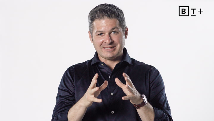 A man in a dark shirt gestures with his hands while speaking, standing in front of a plain white background with a "BT+" logo in the top right corner.