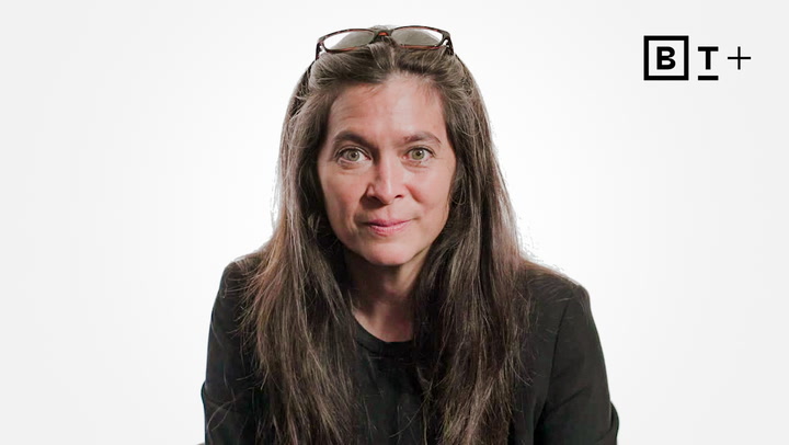 A woman with long brown hair and glasses on her head wears a black top, sitting against a plain white background with a "BT+" logo in the corner.