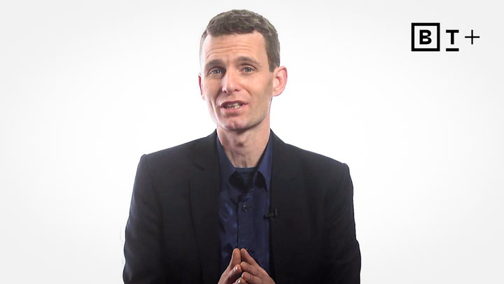 A man in a dark suit sits against a plain white background, speaking and gesturing with his hands. A "B T+" logo appears in the top right corner.