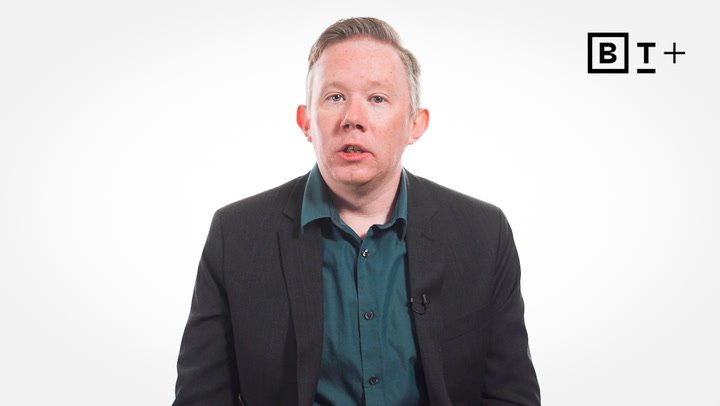 A person in a dark suit and teal shirt sits against a plain white background with a "BT+" logo in the top right corner.