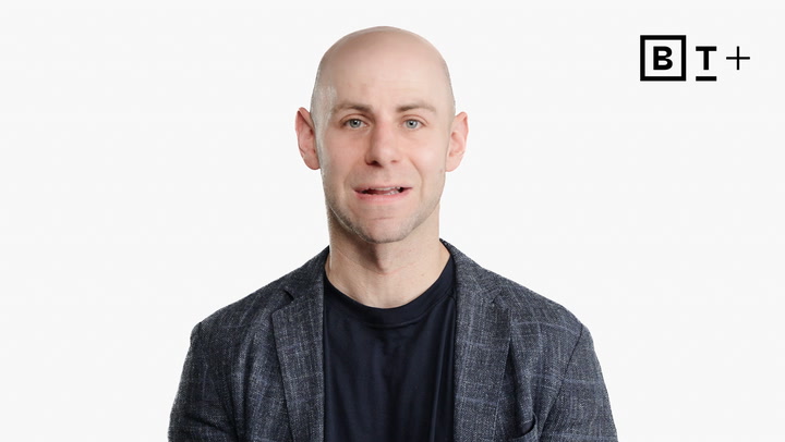 A bald man in a dark shirt and plaid jacket faces the camera in front of a plain white background with the B+ logo in the top right corner.