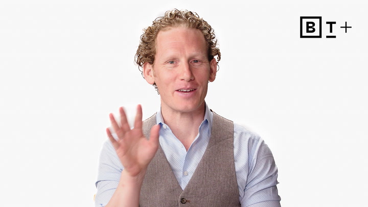 A man with curly hair wearing a light blue shirt and brown vest raises his hand in greeting; a "BT+" logo is visible in the top right corner.