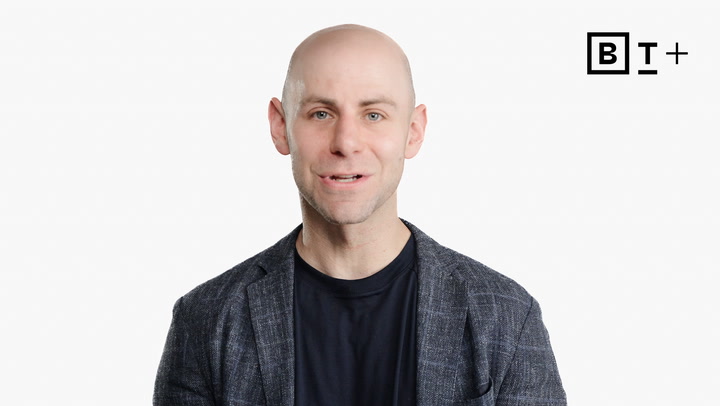 A bald man wearing a dark shirt and checked blazer stands in front of a white background with a "BT+" logo in the top right corner.