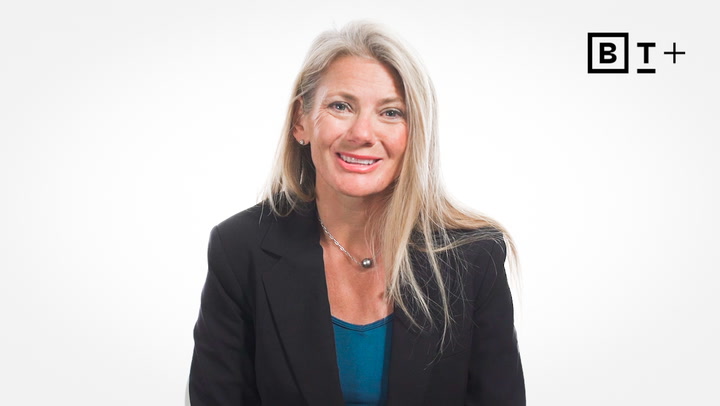 A woman with long blonde hair wearing a black blazer and blue top sits and smiles in front of a plain white background with a "BT+" logo in the top right corner.