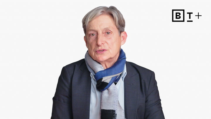 An older person with short gray hair, wearing a dark blazer and a blue-gray scarf, sits in front of a white background with a "B T +" logo in the top right corner.