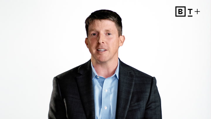 A man in a dark suit jacket and light blue shirt sits against a plain white background with a "BT+" logo in the top right corner.
