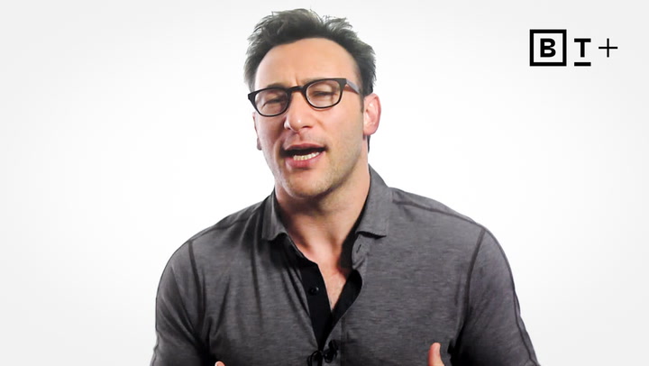 A man wearing glasses and a grey shirt speaks while gesturing with his hands, set against a plain white background with a logo in the top right corner.