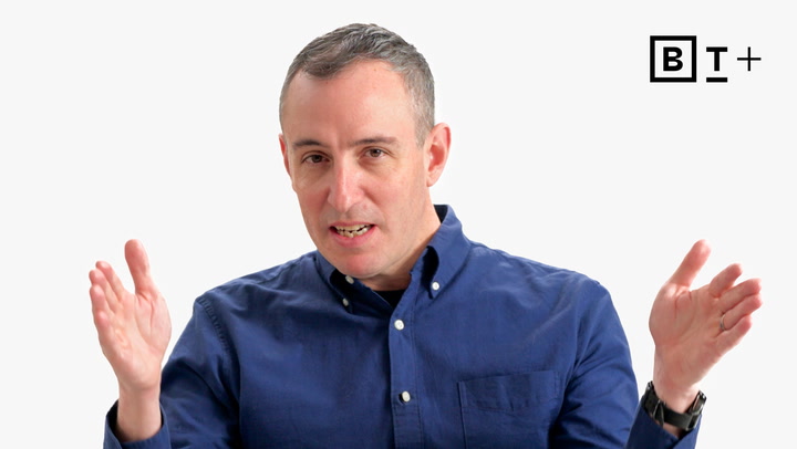 A man in a blue button-up shirt gestures with both hands while speaking, with a "B T +" logo in the top right corner.